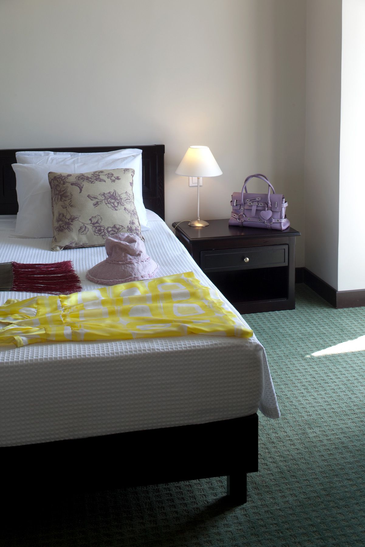 Una cama bien hecha con un cojín floral, una colcha amarilla a cuadros y una manta con flecos rojos. Hay una mesita de noche con una lámpara y un bolso púrpura. La habitación tiene una alfombra verde y paredes blancas.