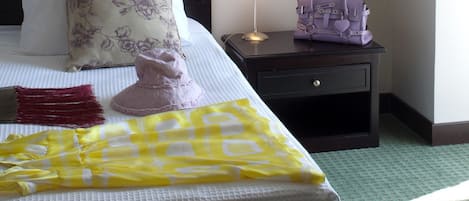 A neatly made bed with a floral pillow, a yellow checkered blanket, and a red fringe throw. There is a nightstand with a lamp and a purple handbag. The room has a green carpet and white walls.