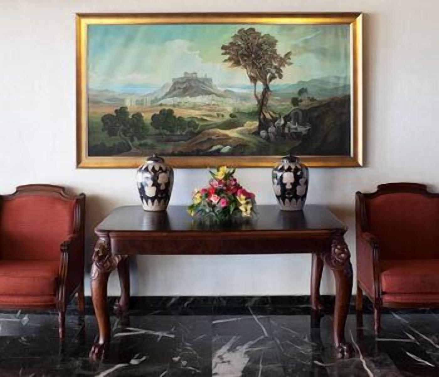 A room with a marble floor, a painting of a landscape, two vases with floral arrangements, and two red upholstered chairs.