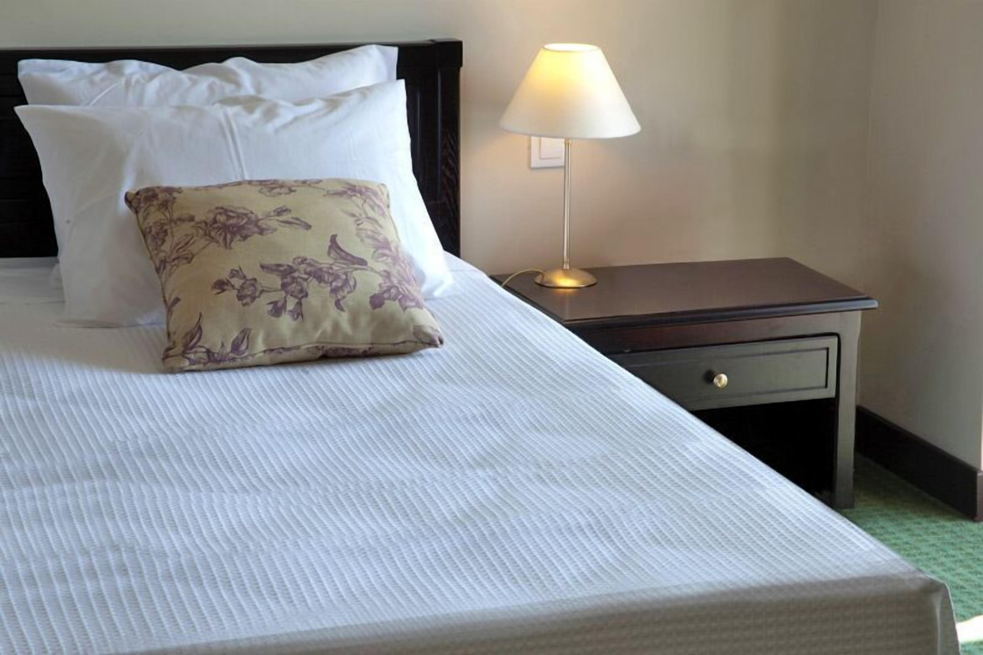 A neatly made bed with white and floral pillows, a bedside table with a lamp, and a drawer.