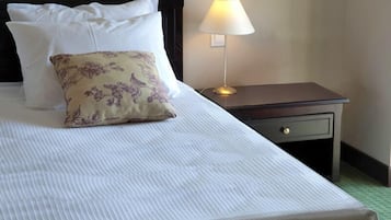 A neatly made bed with white and floral pillows, a bedside table with a lamp, and a drawer.