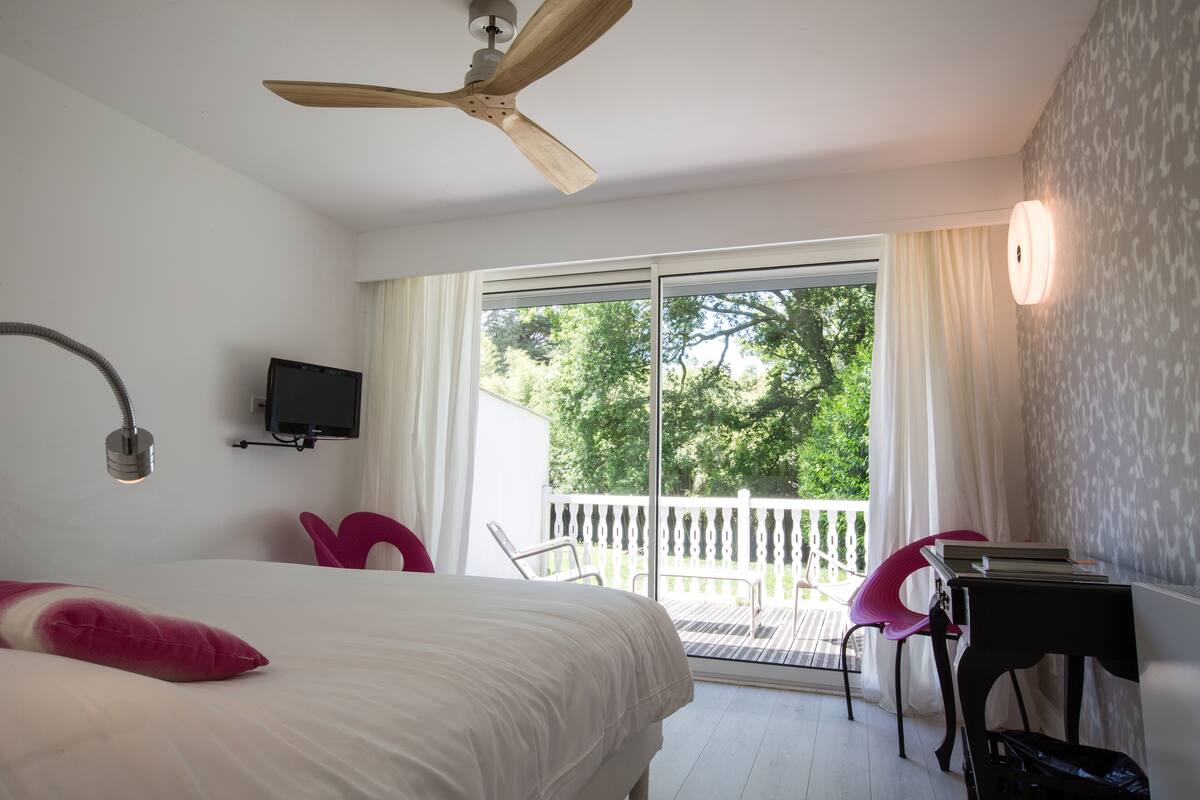 Een slaapkamer met een bed, een plafondventilator, een televisie, een balkon met stoelen en een tafel met boeken.