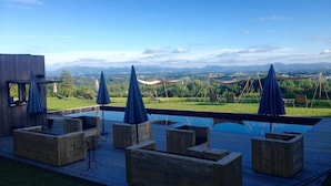 Un espace piscine doté d’une terrasse en bois avec des sièges et des parasols, offrant une vue panoramique sur un paysage magnifique, avec des montagnes à l’horizon.