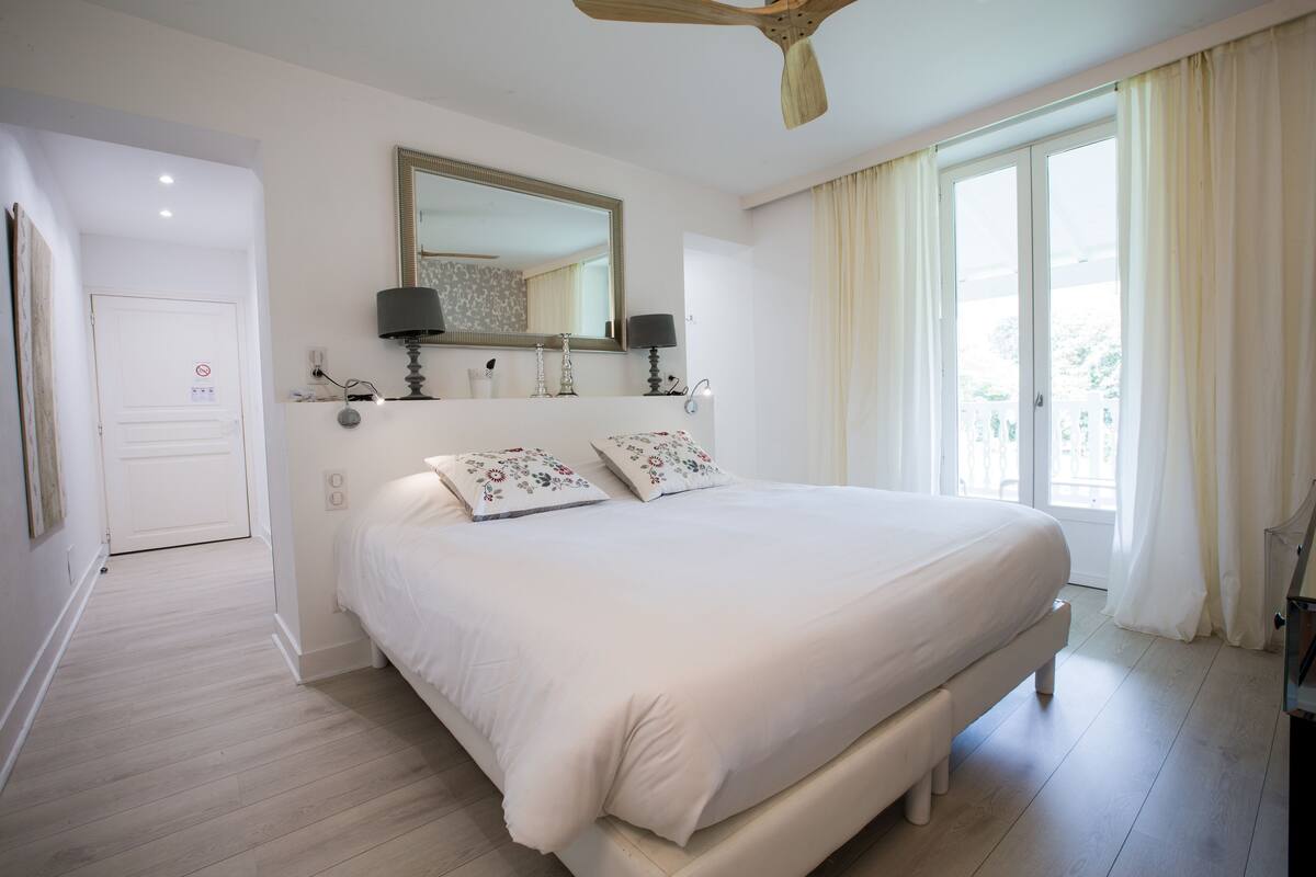 Een slaapkamer met een groot bed, een plafondventilator, een raam met gordijnen en een deur.
