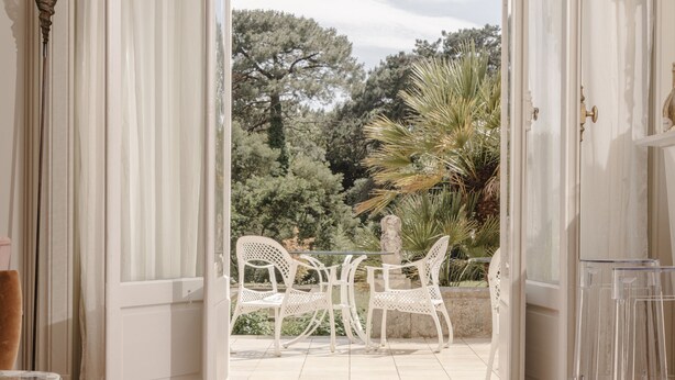 Une porte ouverte donne sur un balcon où se trouvent des chaises blanches, et d’où l’on peut admirer la verdure environnante.