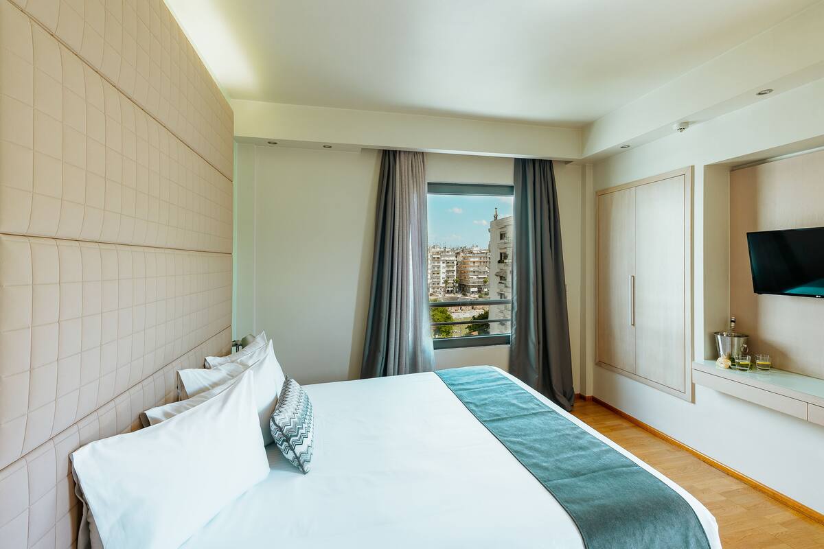 Habitación de hotel con cama, televisor, ventana con cortinas y vista a edificios.