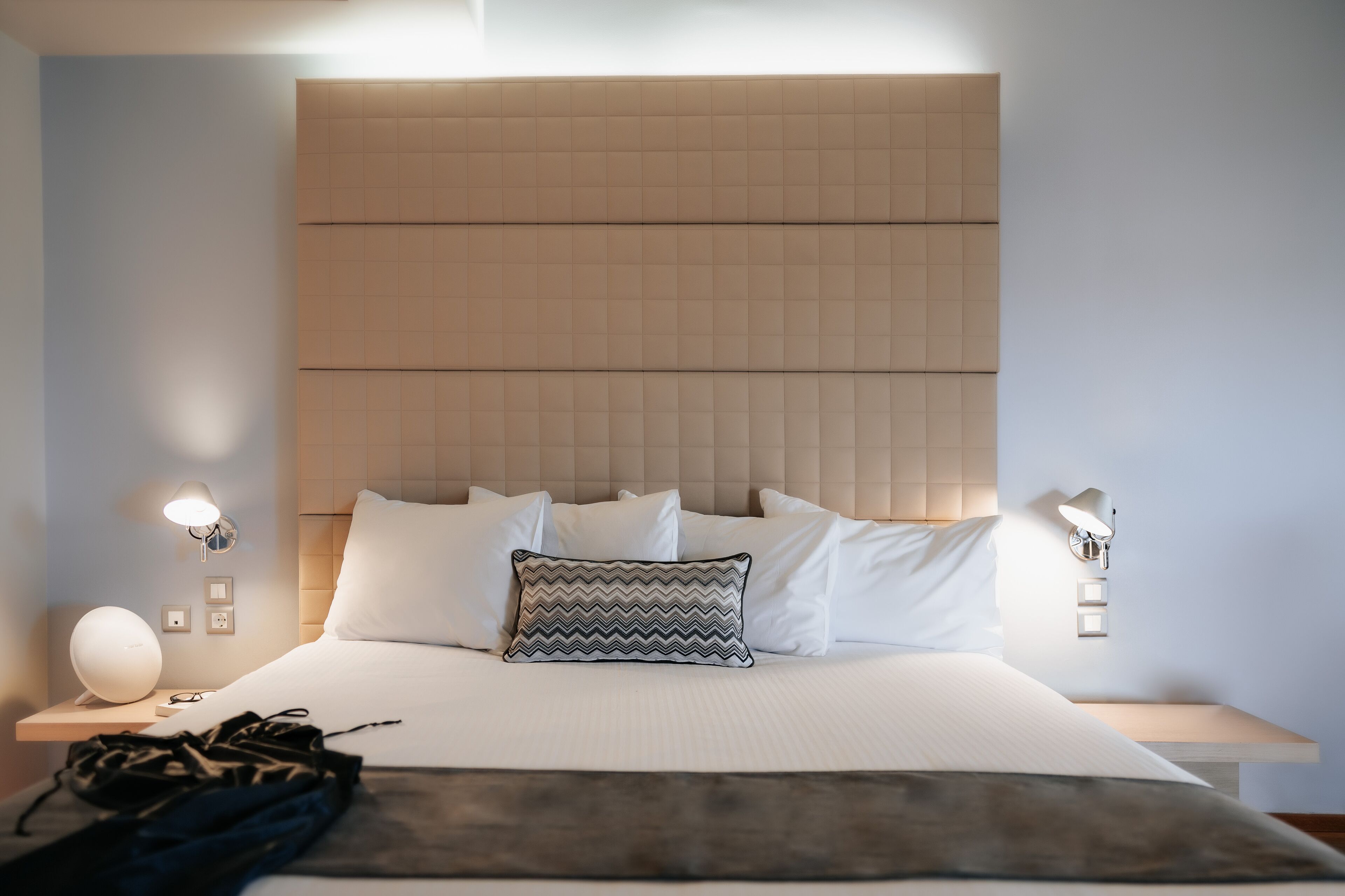 Ein ordentlich bezogenes Bett mit weißen Leinen, einem gemusterten Kopfkissen und einem Nachttisch mit Lampe und einem kleinen Lautsprecher.