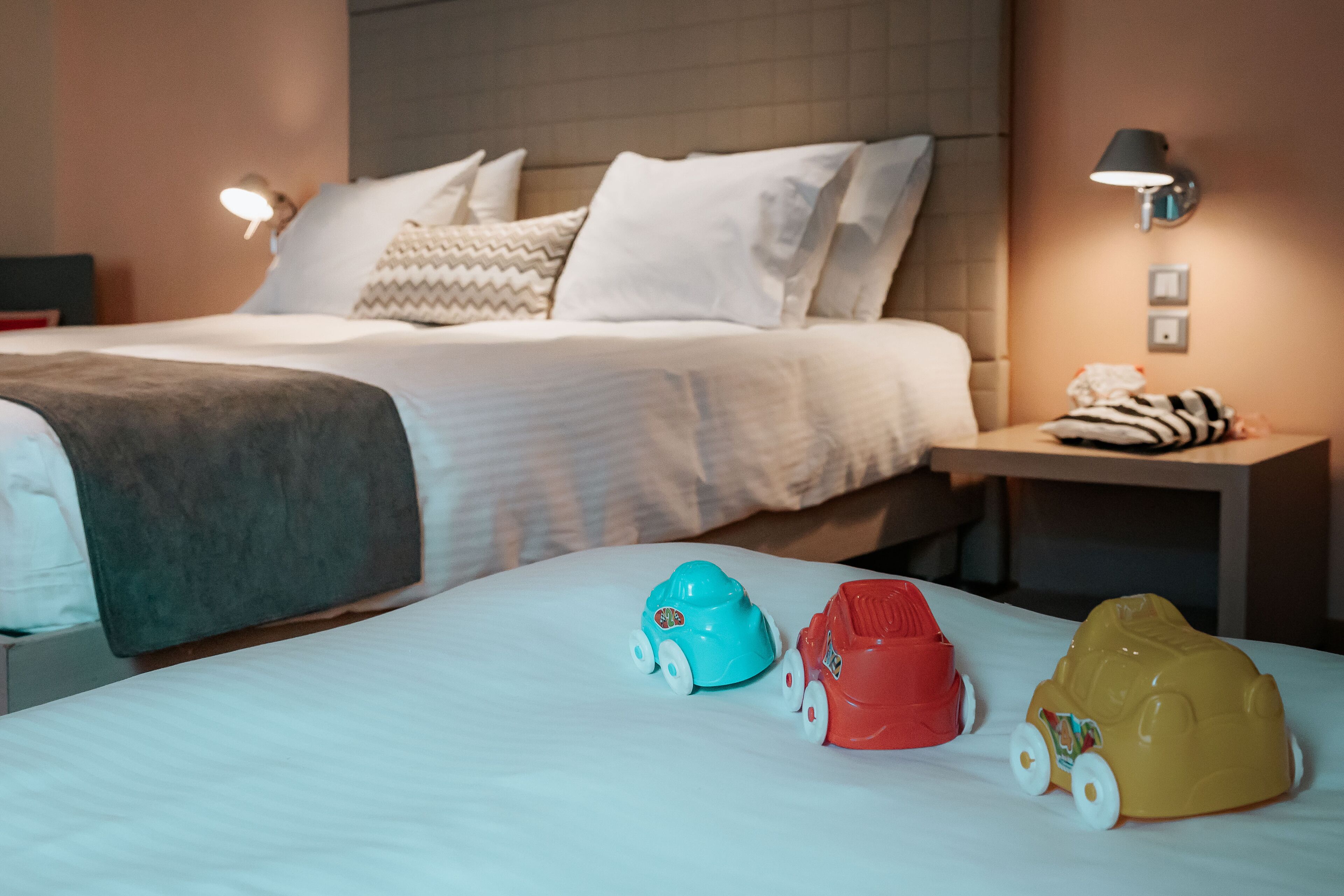 Habitación de hotel con una cama bien hecha, una mesita de noche con lámpara y tres coches de juguete sobre la cama.
