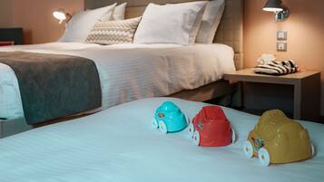 Habitación de hotel con una cama bien hecha, una mesita de noche con lámpara y tres coches de juguete sobre la cama.