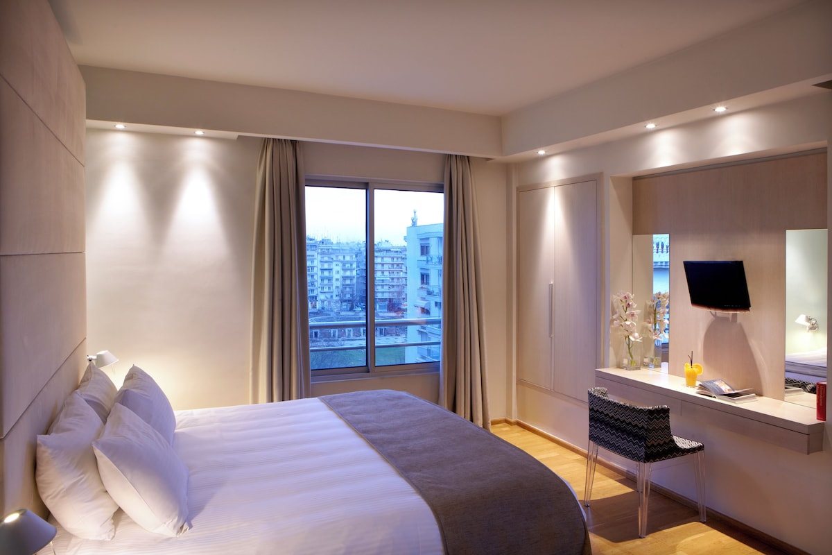 Une chambre d’hôtel moderne dotée d’un grand lit, d’un bureau avec un ordinateur et offrant une vue sur la ville grâce à une grande fenêtre.