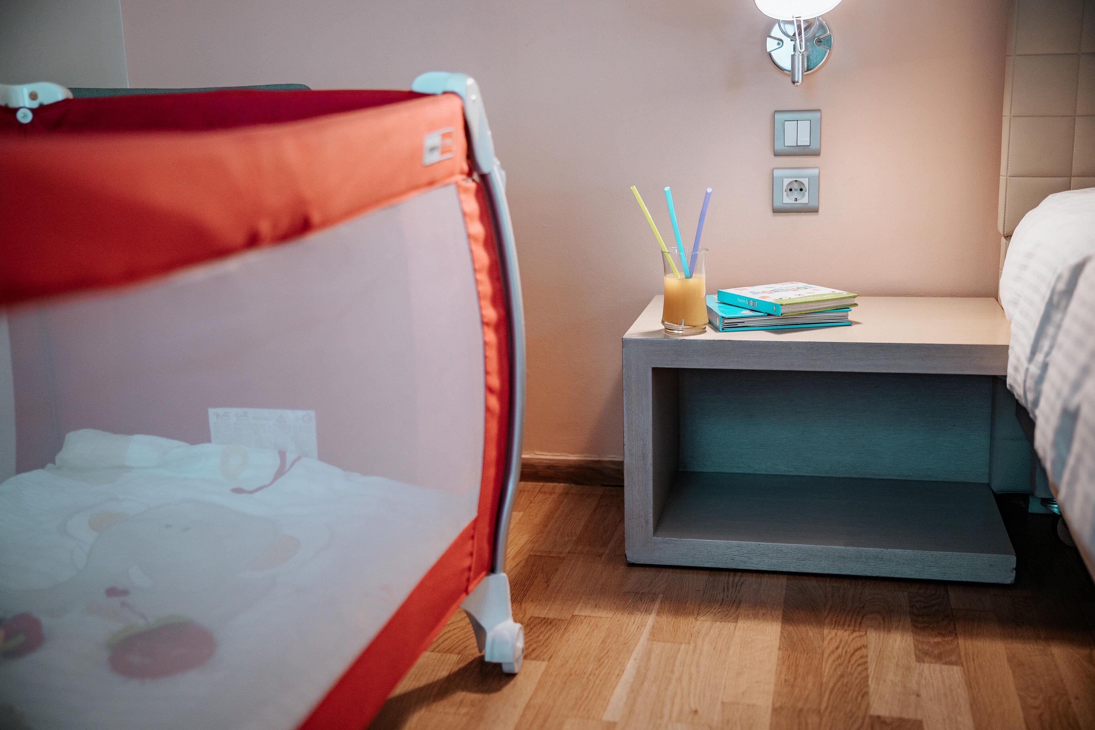 A child's playpen with a white mattress and a red canopy, a bedside table with a lamp, a wall socket, and a stack of books.