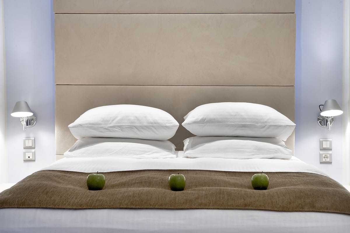 Una cama bien hecha con sábanas blancas, dos almohadas y una manta marrón. Hay tres manzanas verdes sobre la manta.
