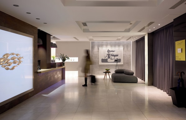 En moderne hotellobby med reception, et stort abstrakt maleri, et siddeområde og en plante.