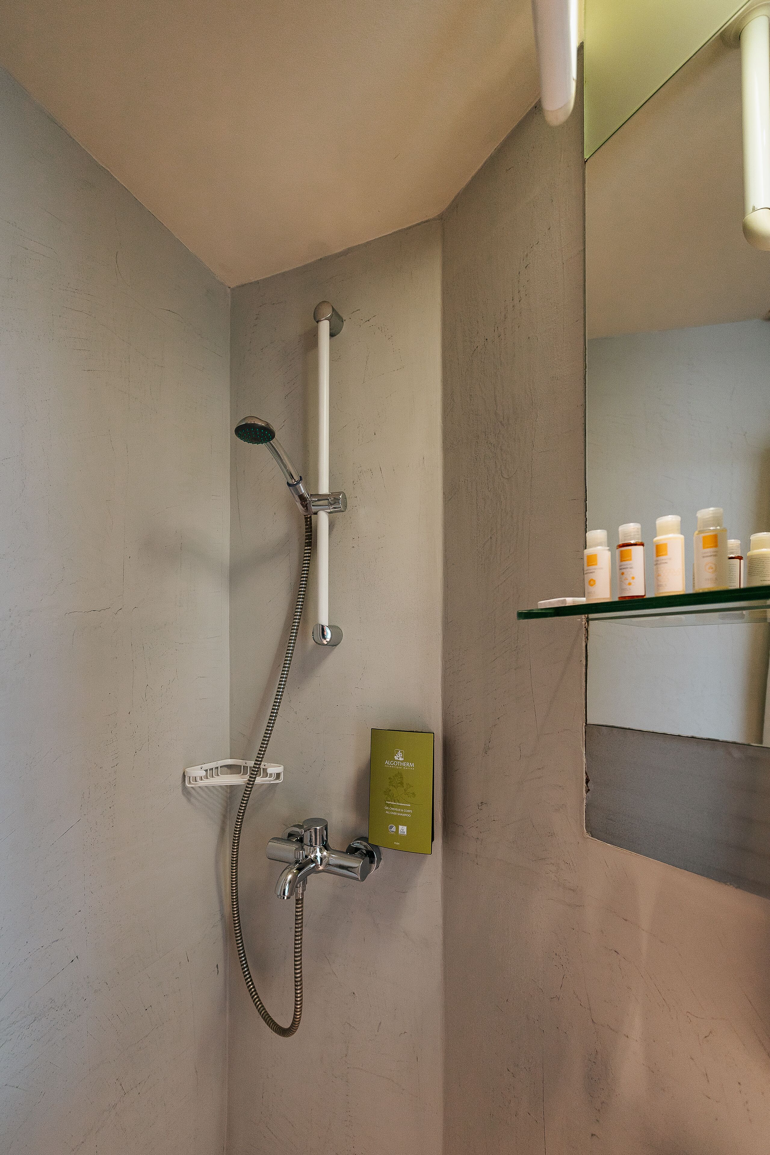 A shower with a handheld showerhead and a wall-mounted faucet. There is a shelf with toiletries.