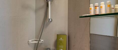 A shower with a handheld showerhead and a wall-mounted faucet. There is a shelf with toiletries.