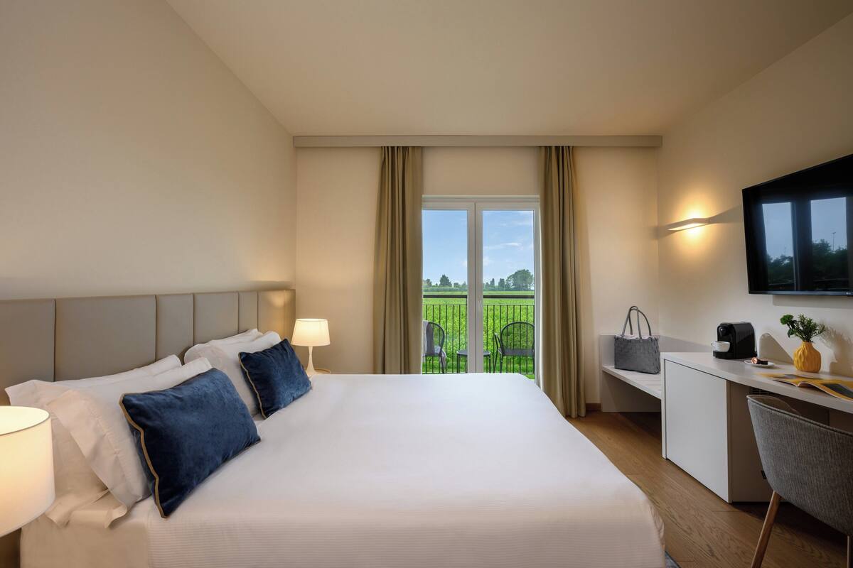 A hotel room with a large bed, a desk, a chair, a TV, and a balcony with chairs.