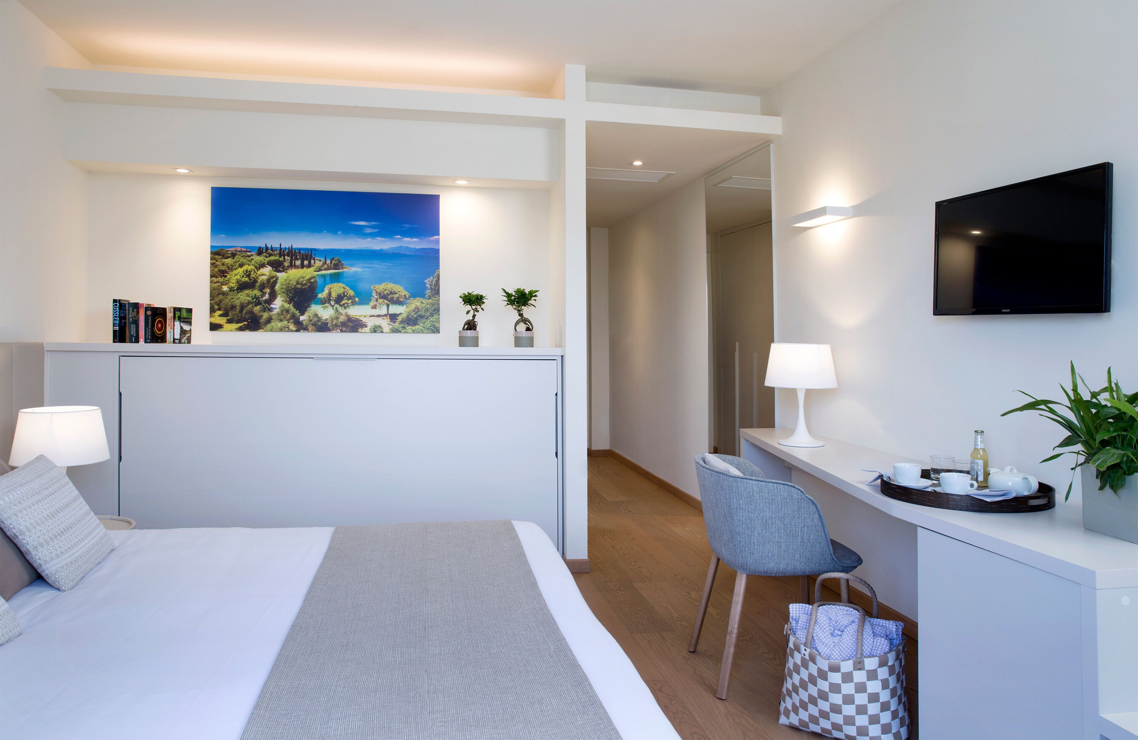 A modern hotel room with a large bed, a desk with a chair, a flat-screen TV, and a painting of a coastal landscape.