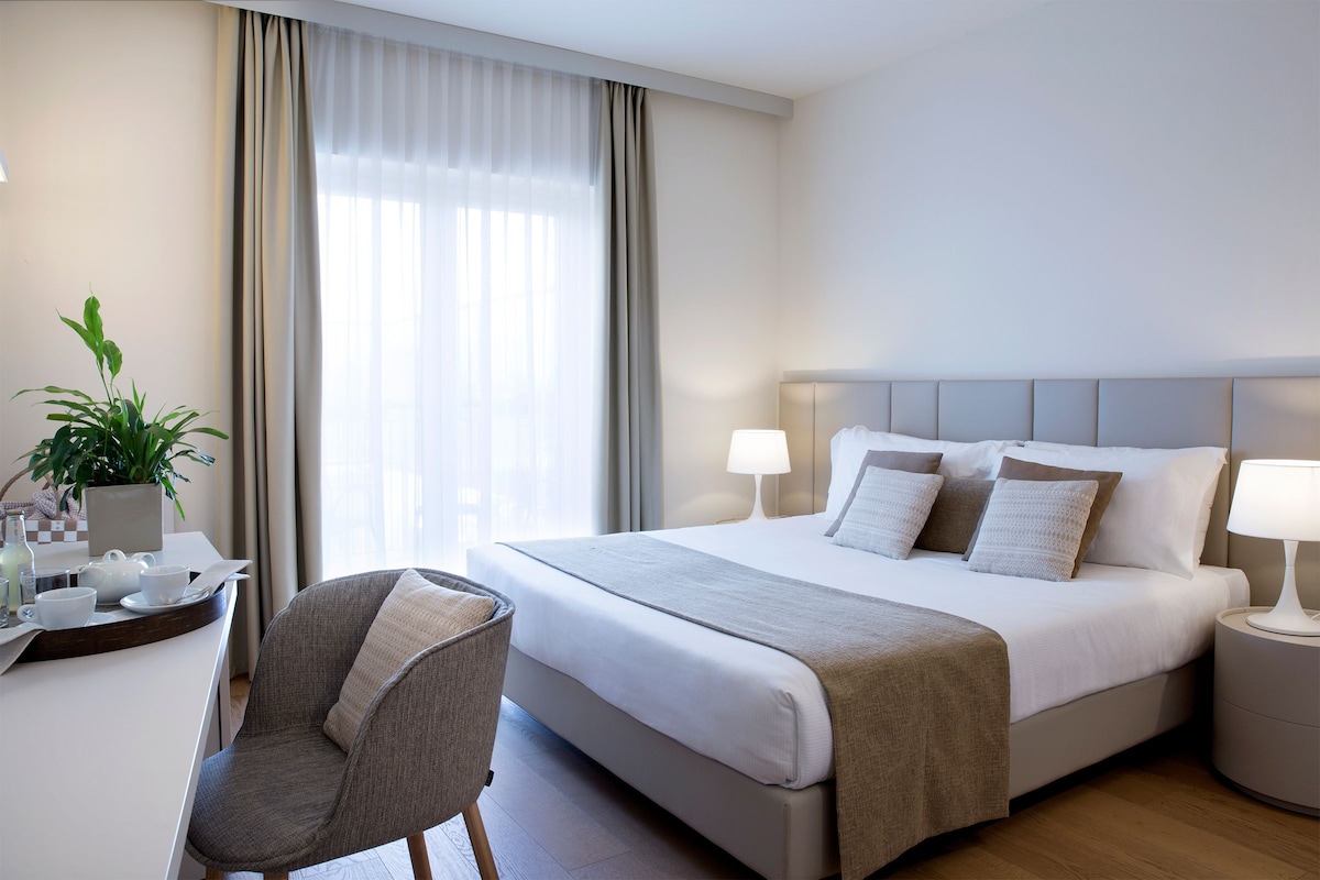 A hotel room with a bed, bedside tables, a chair, a desk with a plant, and a window with curtains.