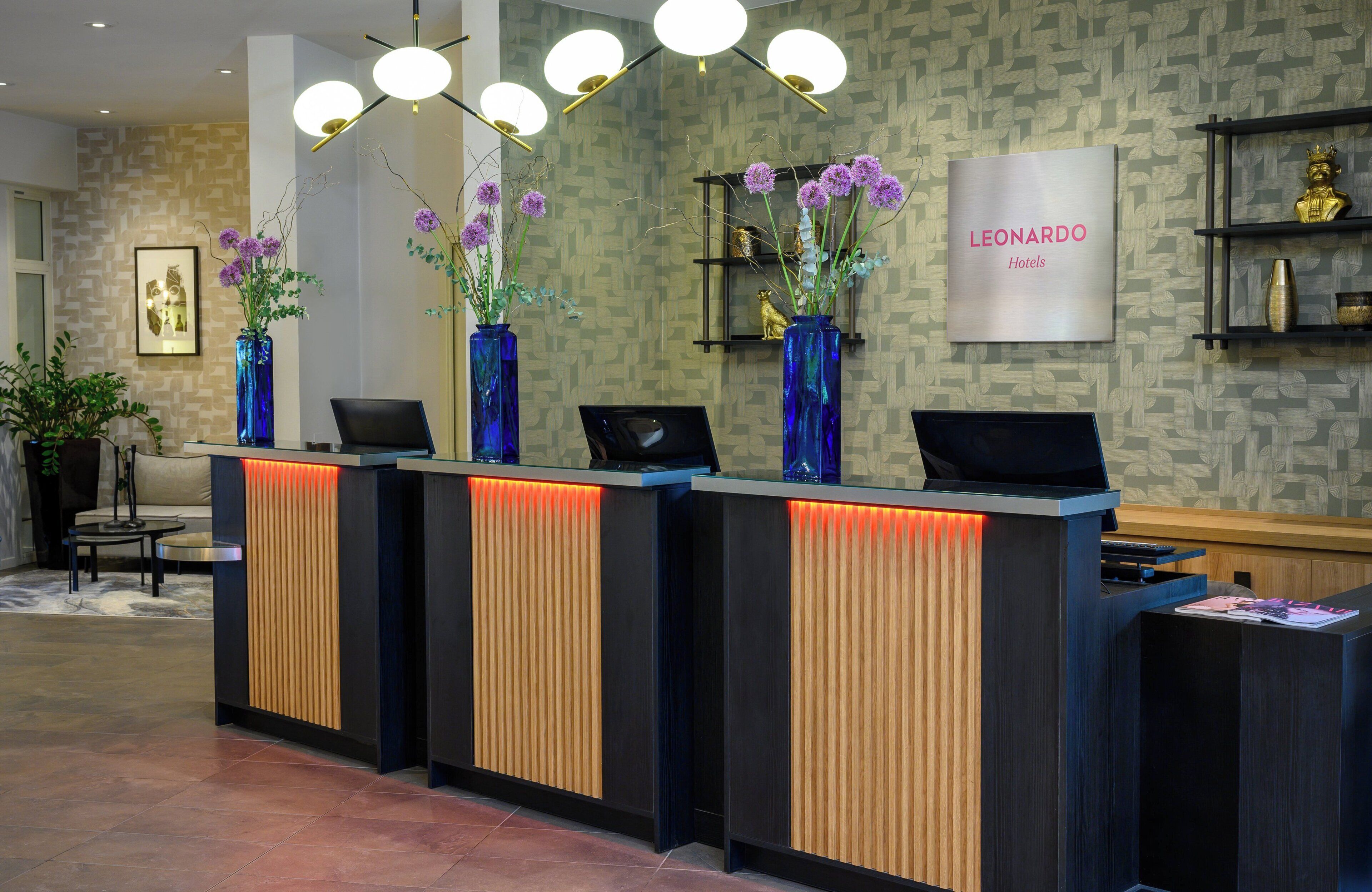 A hotel reception desk with three blue vases of purple flowers, a framed picture on the wall, and a 'LEONARDO Hotels' sign.