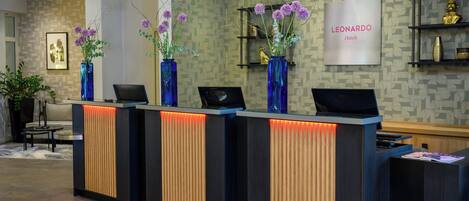 A hotel reception desk with three blue vases of purple flowers, a framed picture on the wall, and a 'LEONARDO Hotels' sign.
