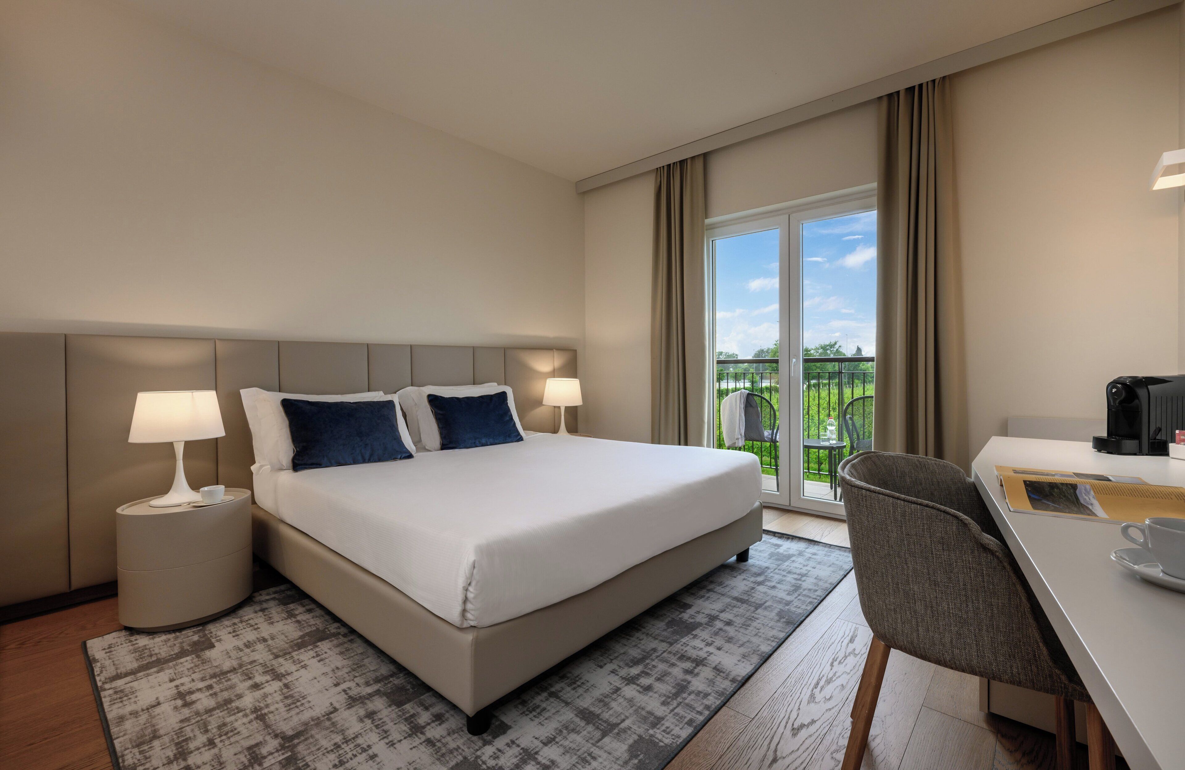 A hotel room with a bed, two bedside tables with lamps, a desk with a chair, and a view of a balcony with greenery.