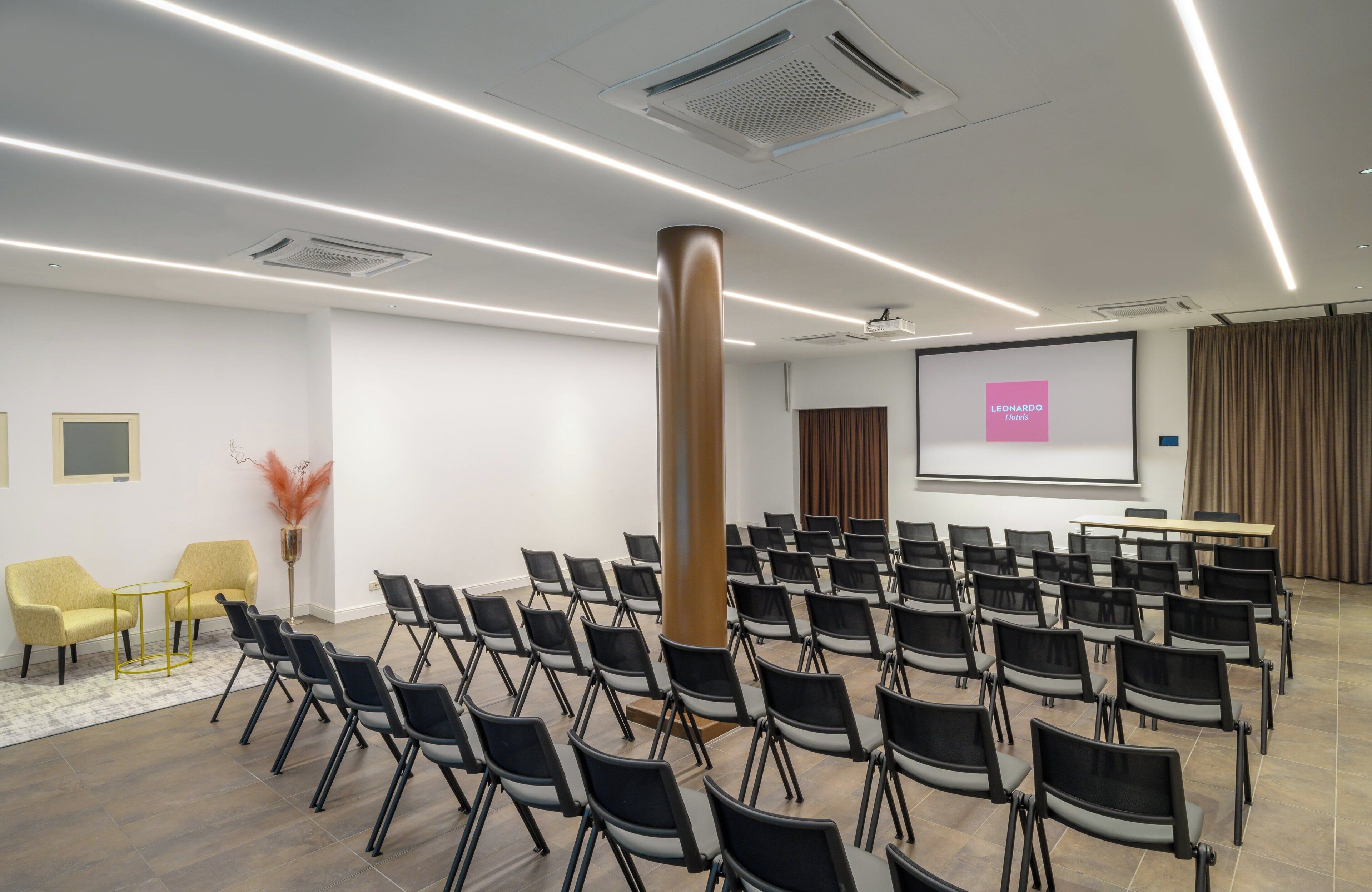 Un salón de conferencias con filas de sillas, un pilar grande, una pantalla de proyector y una pantalla para presentaciones.