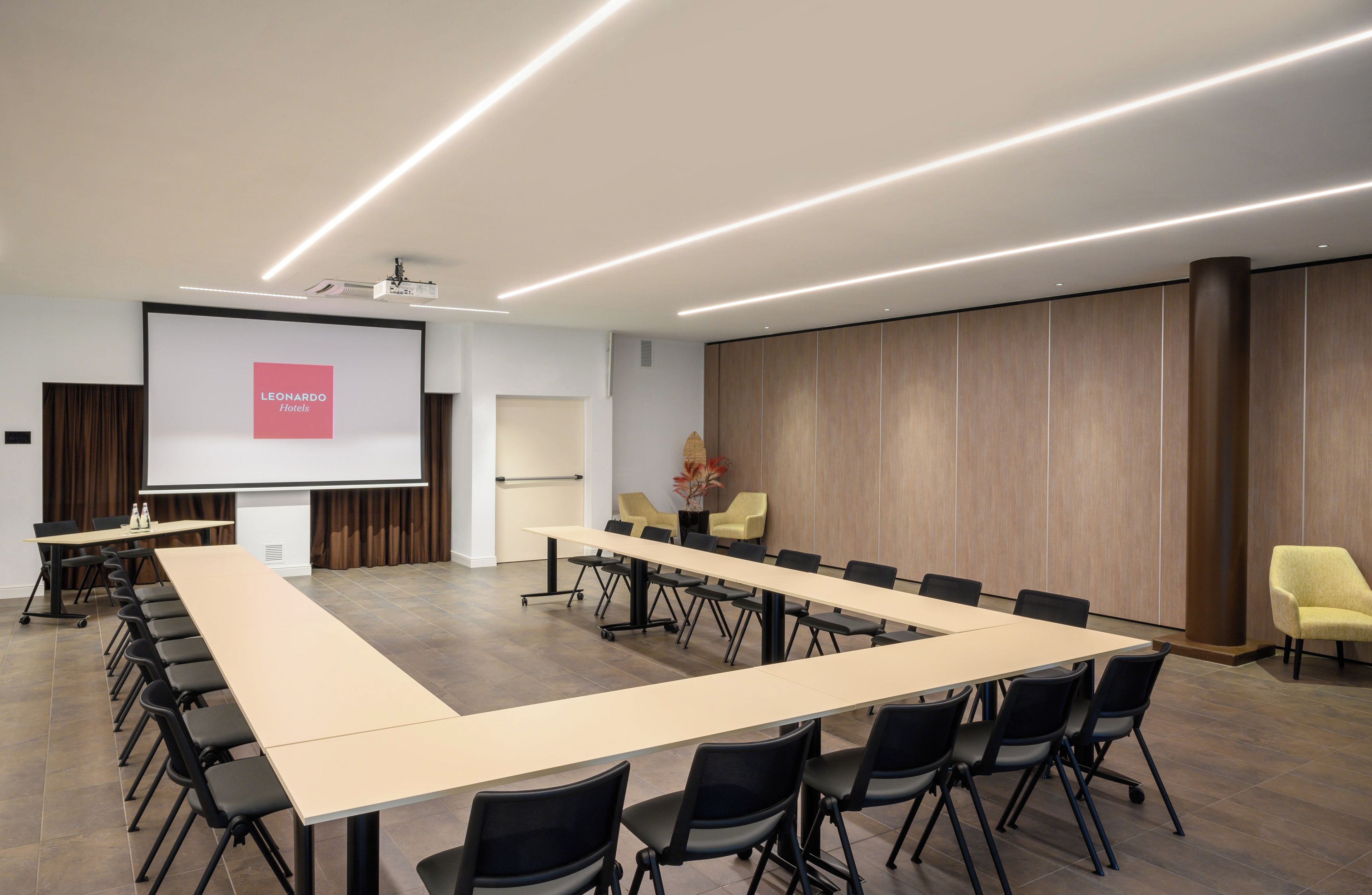 Une salle de conférence avec une longue table, des chaises, un écran de projection et un mur recouvert de panneaux de bois.