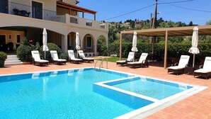 A swimming pool with lounge chairs and umbrellas, surrounded by a building with a balcony and a view of the hillside.