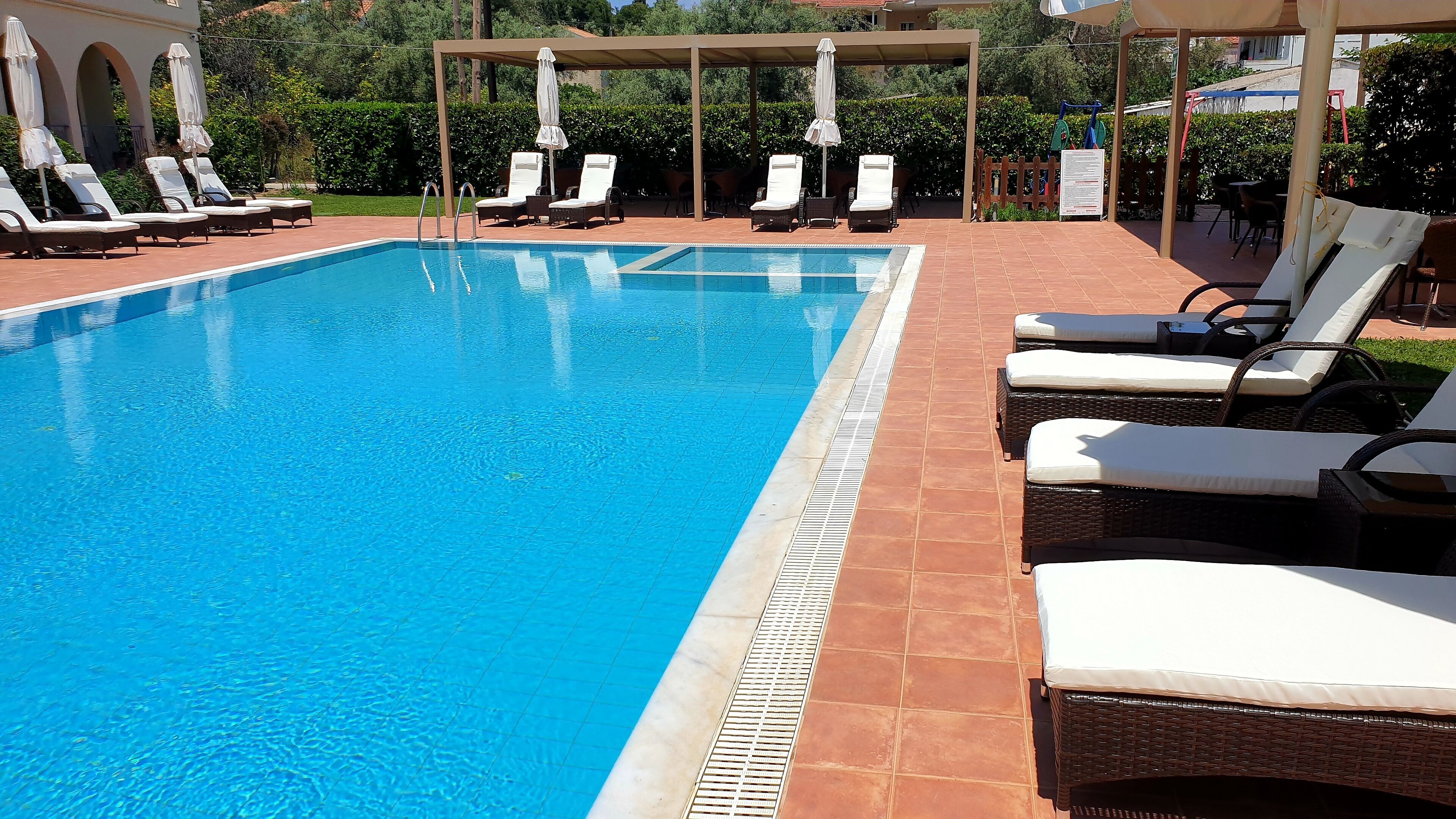 Uma piscina com espreguiçadeiras e guarda-sóis, rodeada por um pátio ladrilhado e vegetação.