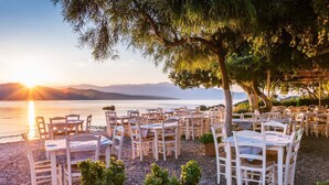 An outdoor dining area with tables and chairs arranged on a pebbled surface, overlooking a body of water with mountains in the background.