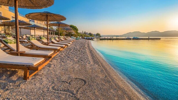 A beach with sun loungers and umbrellas, a pebble beach, and a calm sea with mountains in the background.
