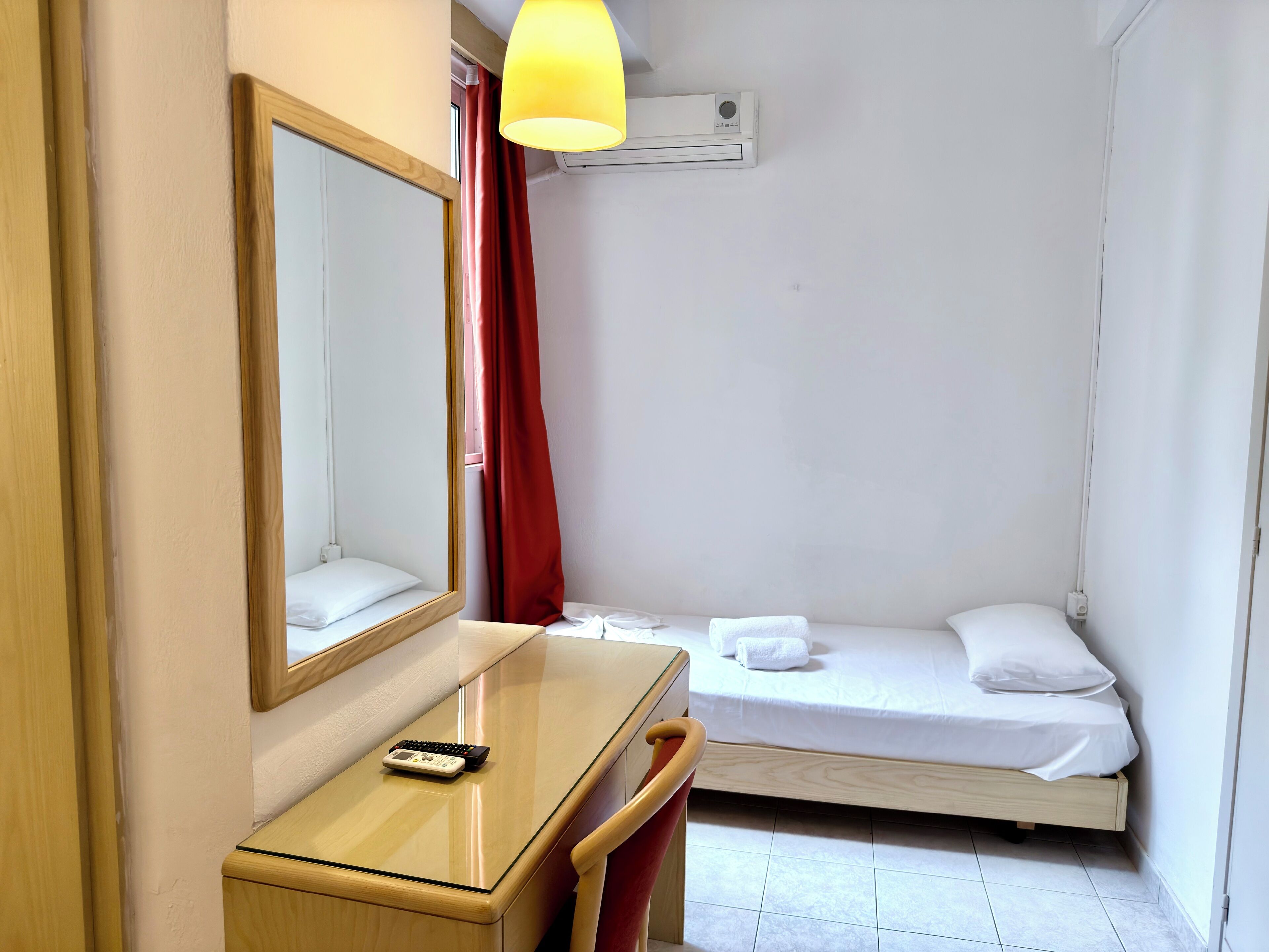 Una habitación pequeña y limpia con cama, mesita de noche con lámpara, espejo y ventana con cortinas rojas.