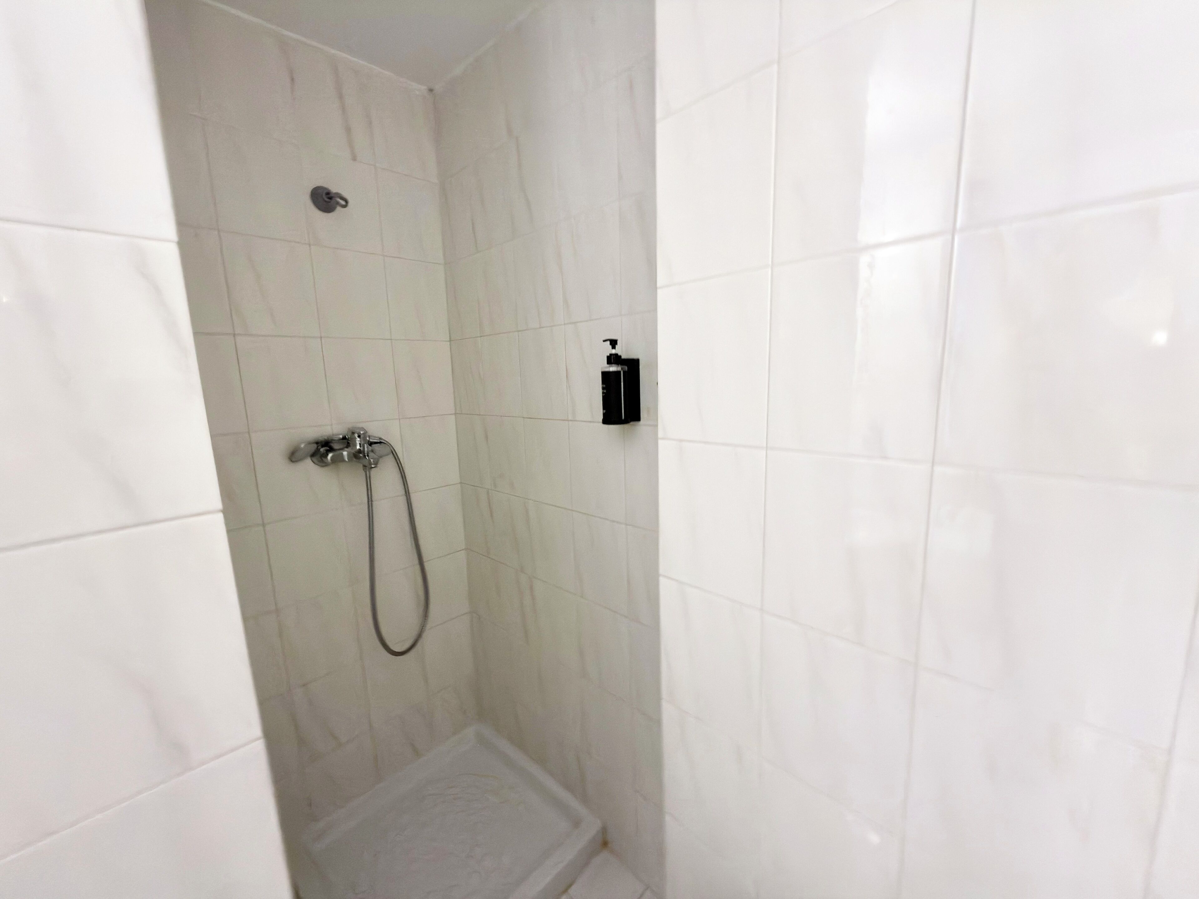 Un pequeño box de ducha con azulejos blancos, un cabezal de ducha manual y dispensador de jabón en la pared.