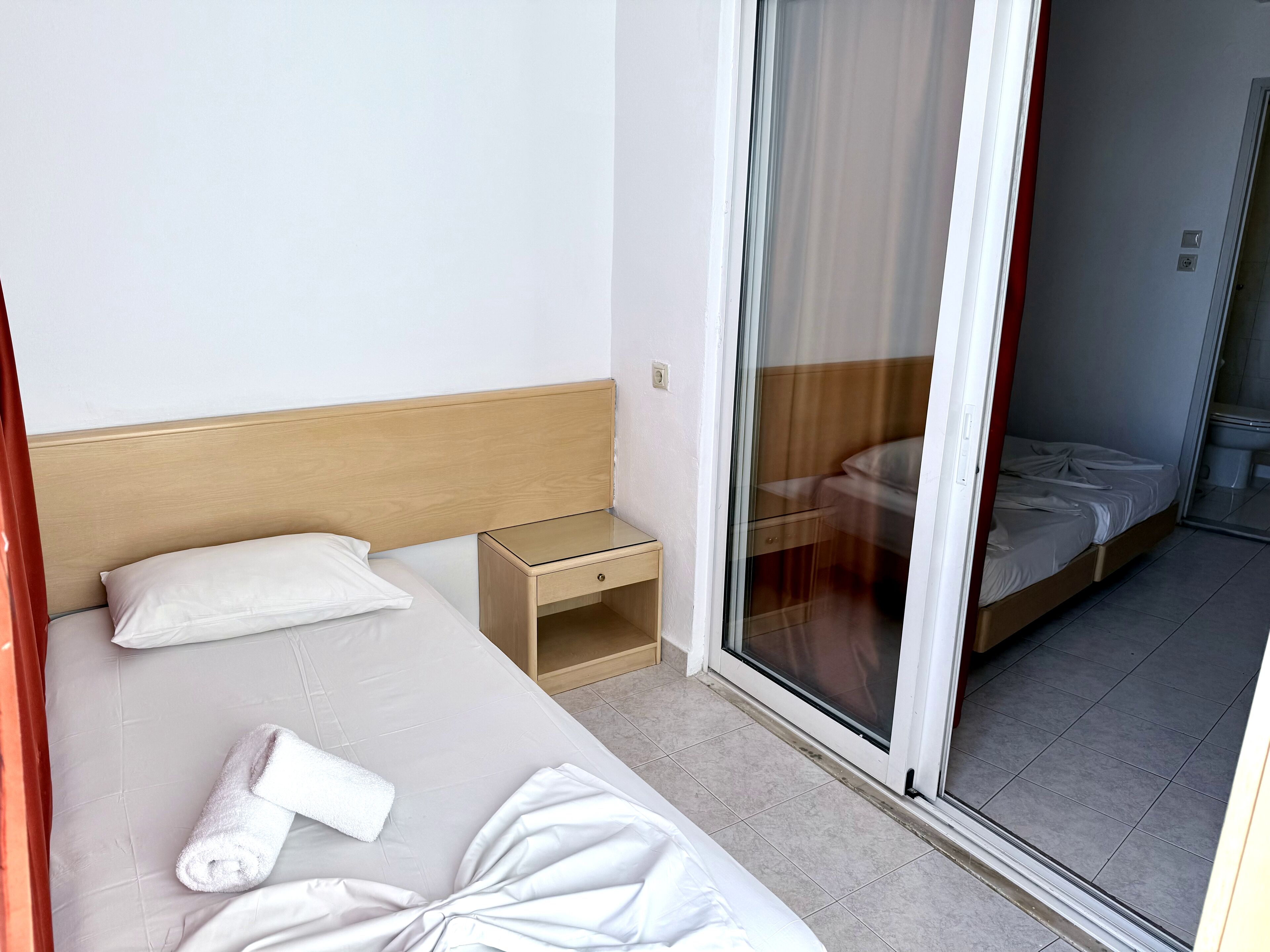 A hotel room with a bed, a small bedside table, a sliding glass door leading to a bathroom, and a view of the outside.