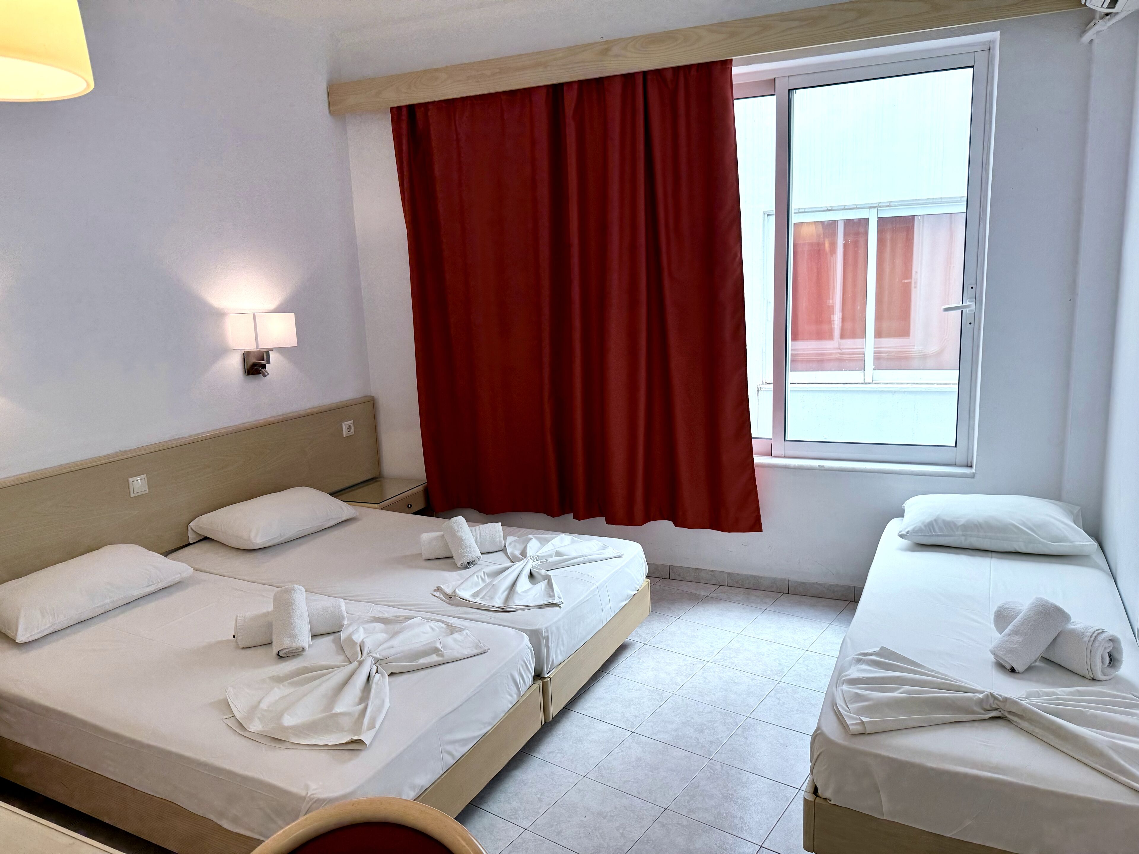 A hotel room with two beds, a window with red curtains, a wall-mounted lamp, and a wooden headboard.