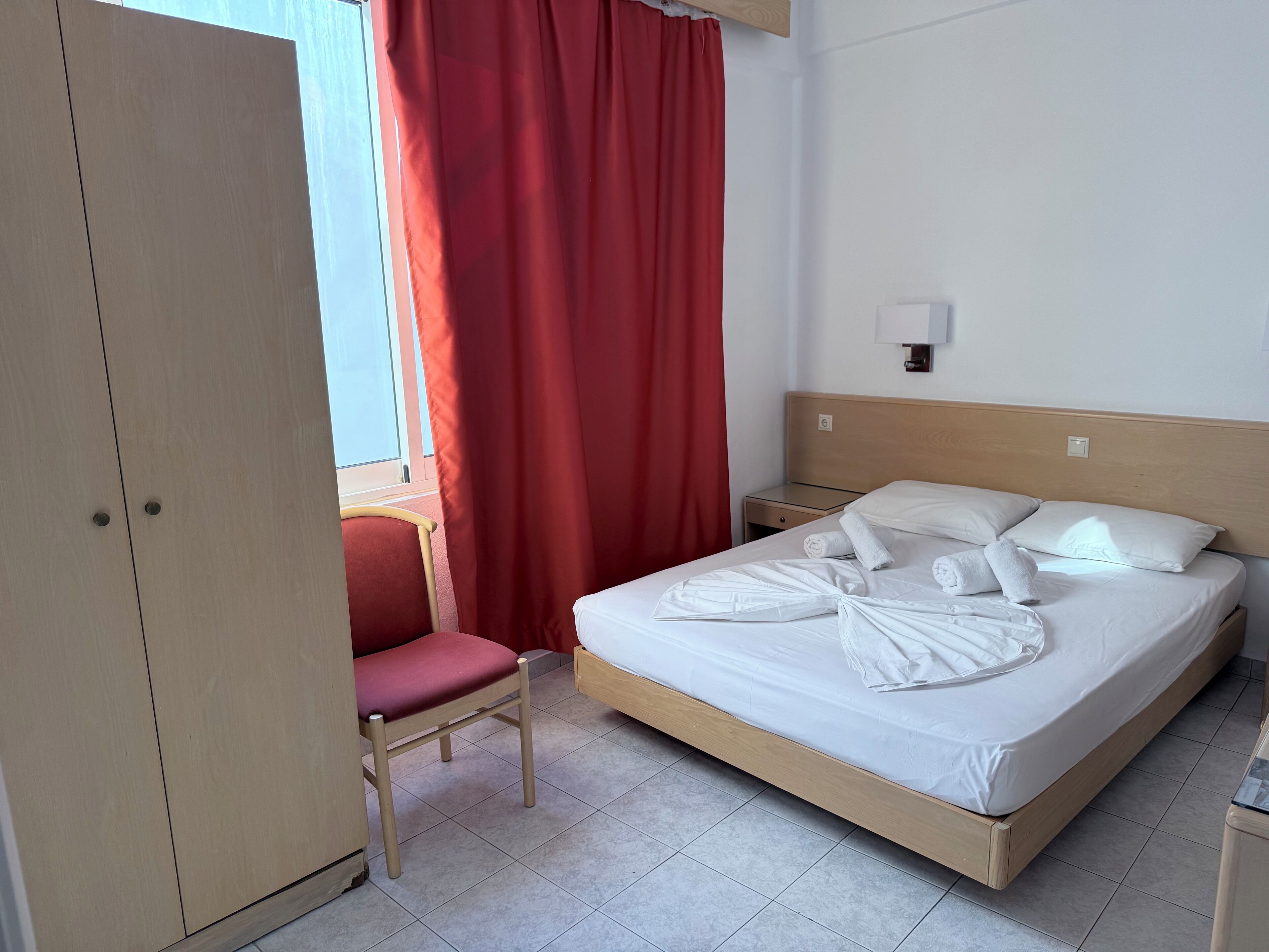 A hotel room with a bed, a chair, a wardrobe, and a window with red curtains.