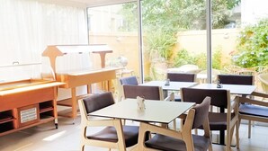 A dining area with tables and chairs, a piano, and a view of the garden through large windows. - Savoy City Hotel (Rhodes)
