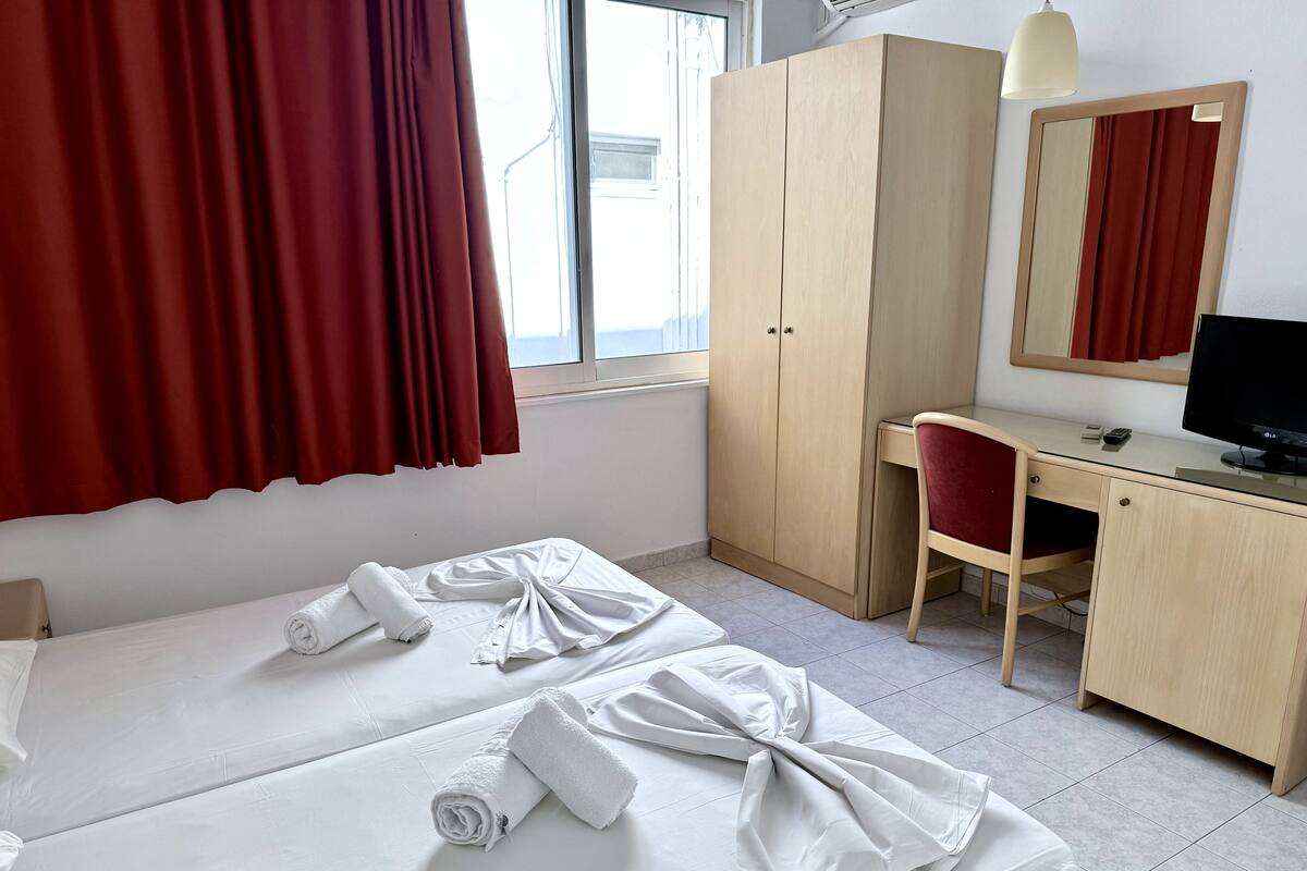 Habitación de hotel con dos camas, un escritorio, una silla, un armario, un televisor y una ventana con cortinas.