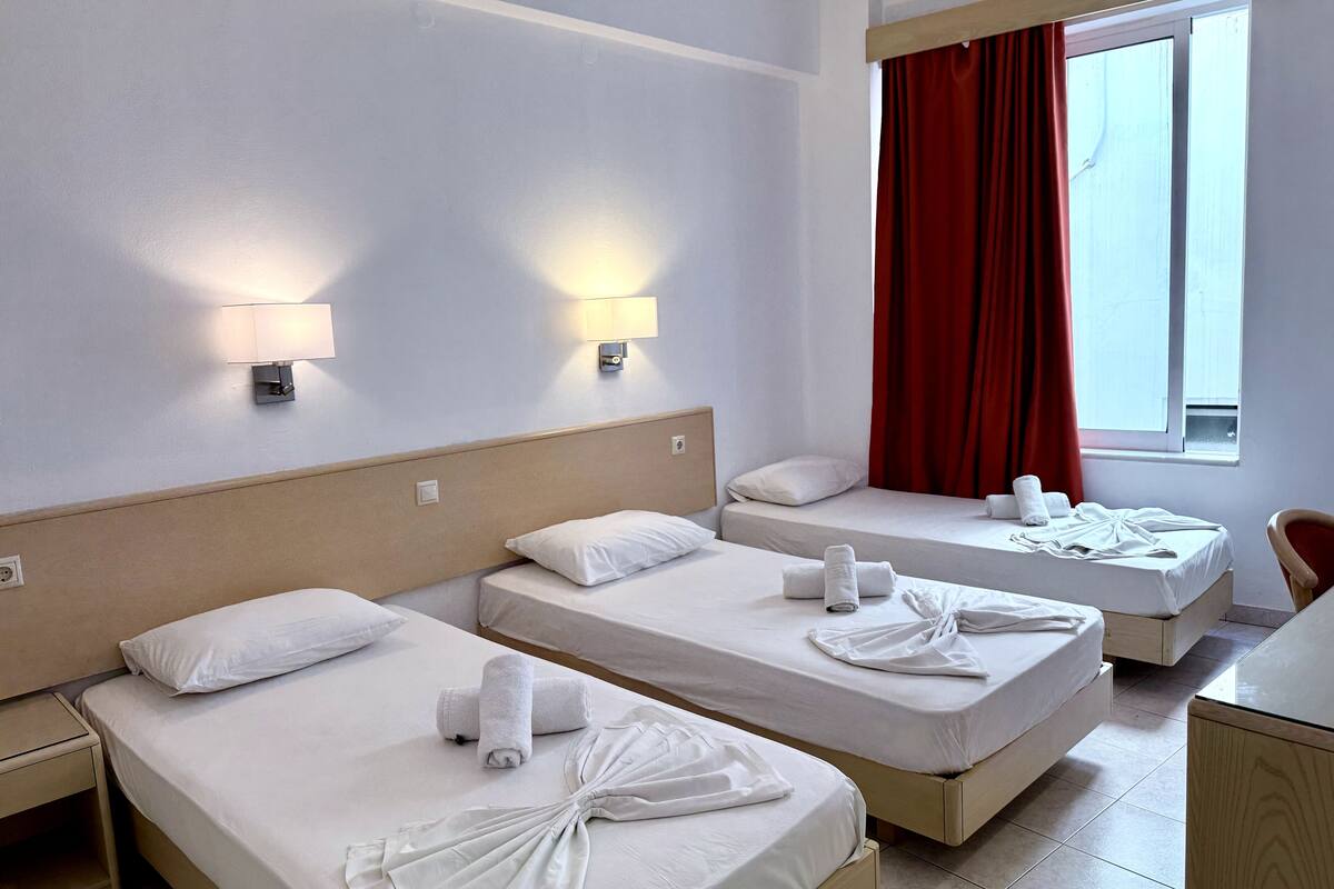 Habitación de hotel con tres camas individuales, cada una con una toalla y una almohada. Hay dos lámparas de pared y un ventanal con cortinas rojas.