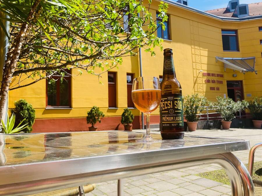 Eine Flasche Pécsi Sör Premium Lager und ein Glas auf einem Metalltisch im Freien, im Hintergrund ein gelbes Gebäude.