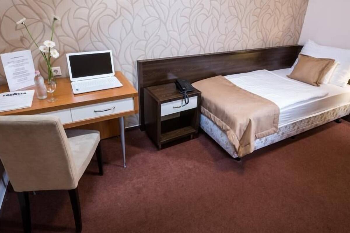 A hotel room with a bed, a desk with a laptop, a chair, a nightstand, and a vase with flowers.