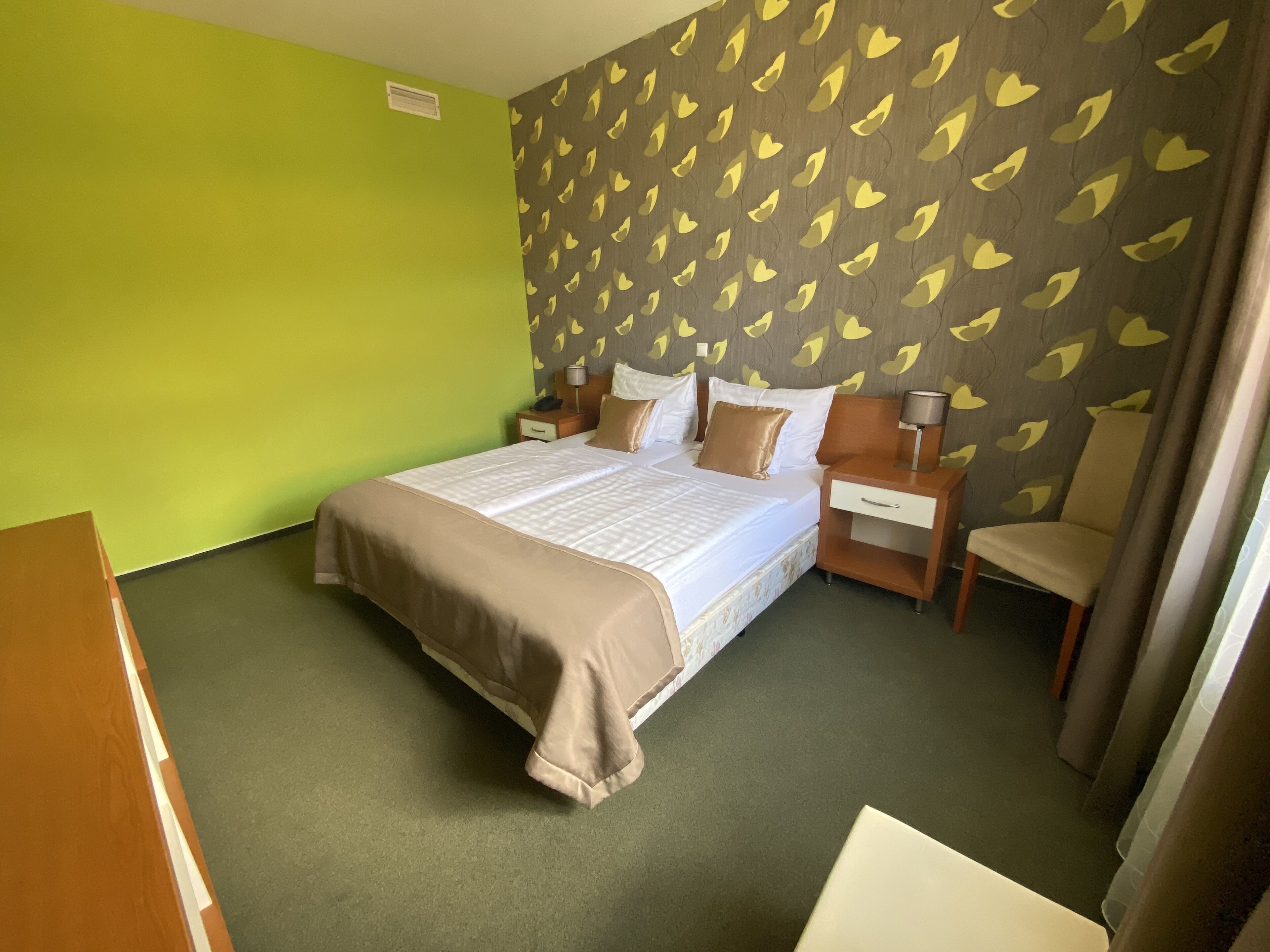 A hotel room with a large bed, two bedside tables with lamps, a chair, and a green wall with a leaf pattern.