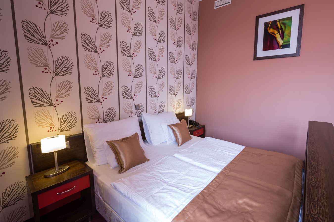 Una cama bien hecha con sábanas blancas y dos almohadas, una mesita de noche con una lámpara, y una pared con estampado floral.
