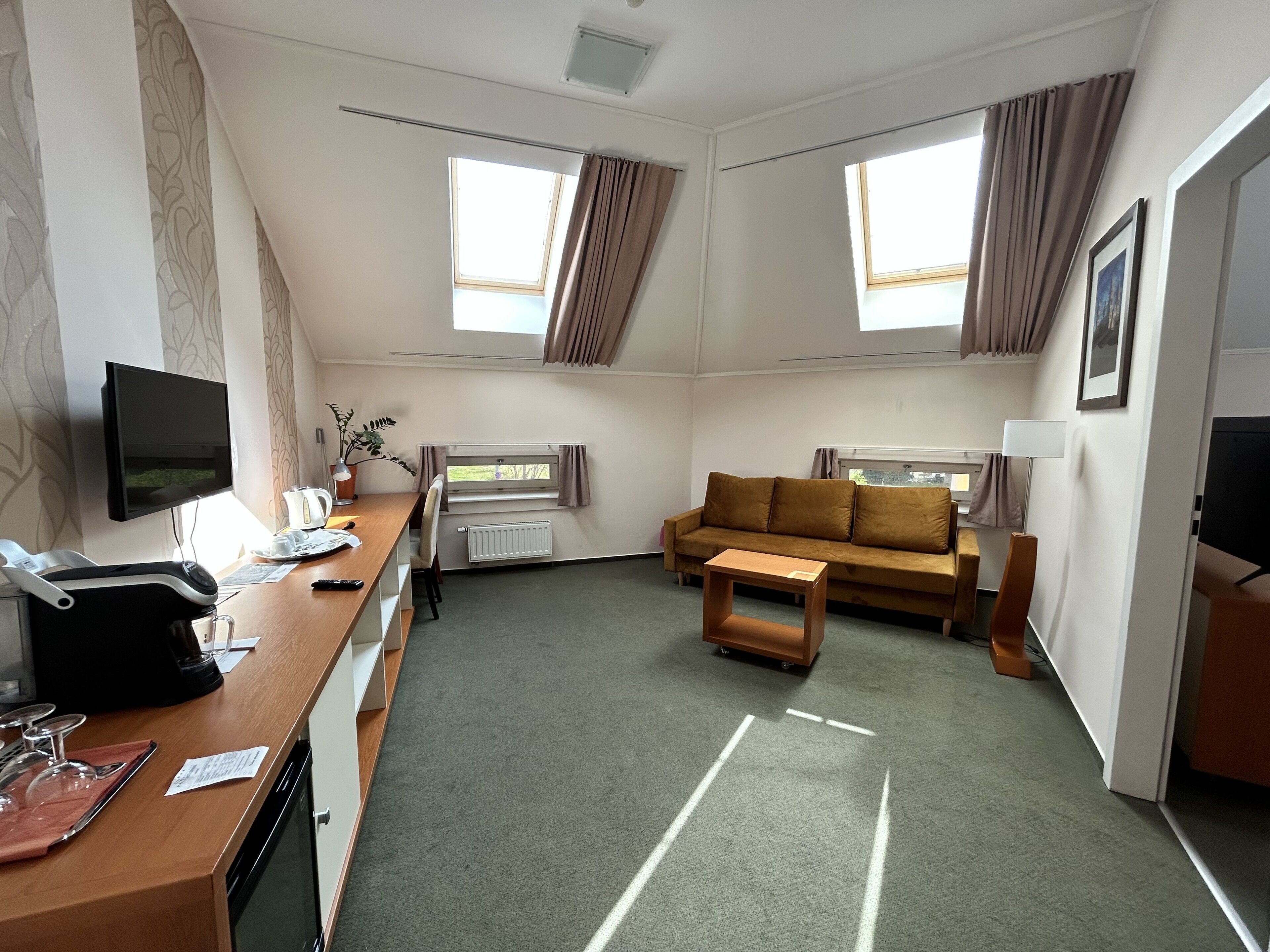 A hotel room with a sofa, a small table, a desk with a computer and a chair, a TV, and two windows with curtains.