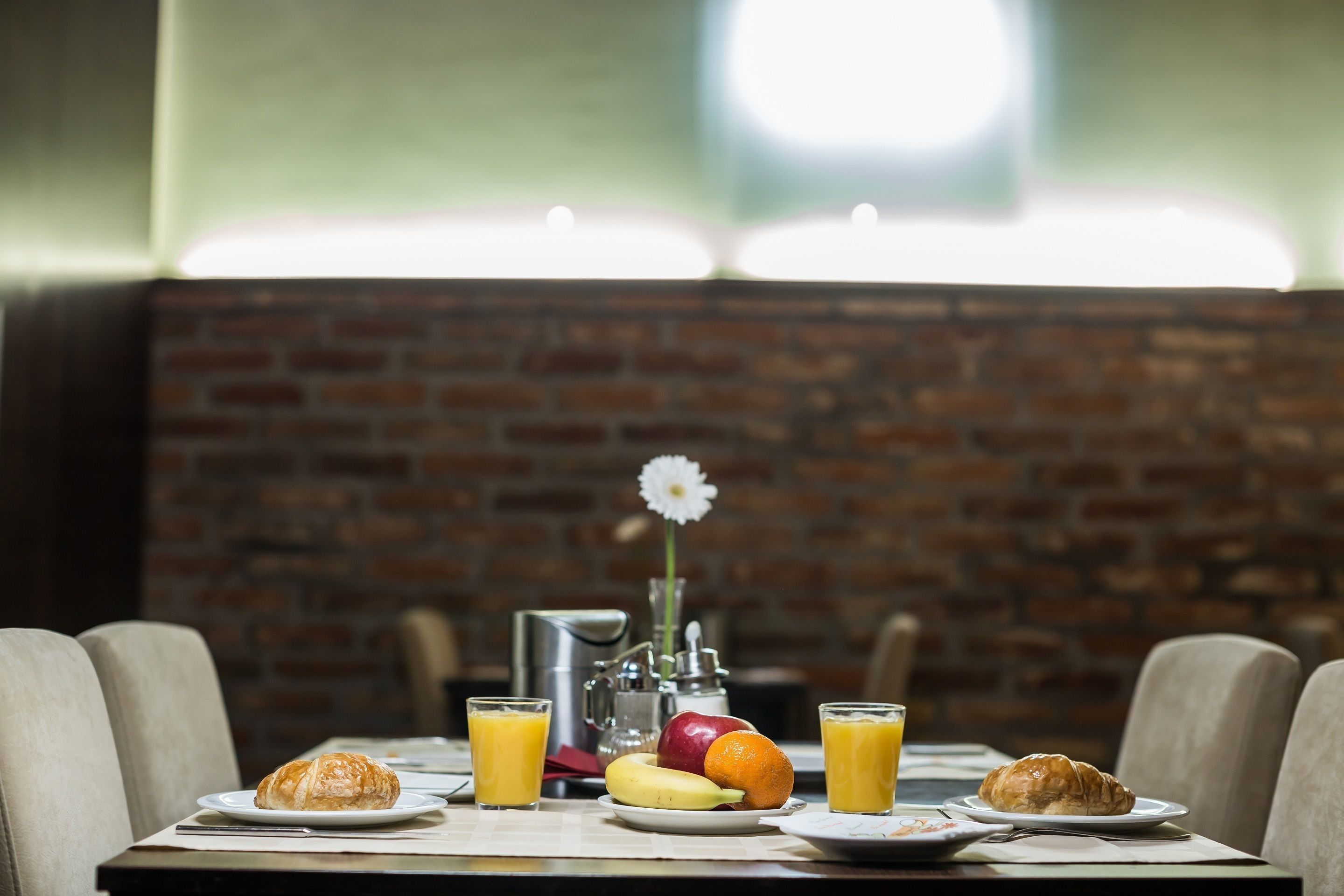 A breakfast setting with croissants, fruit, and orange juice on a table, with a brick wall and a flower in the background.