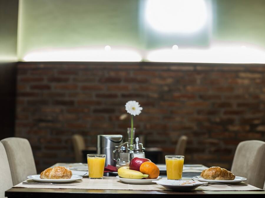 A breakfast setting with croissants, fruit, and orange juice on a table, with a brick wall and a flower in the background.