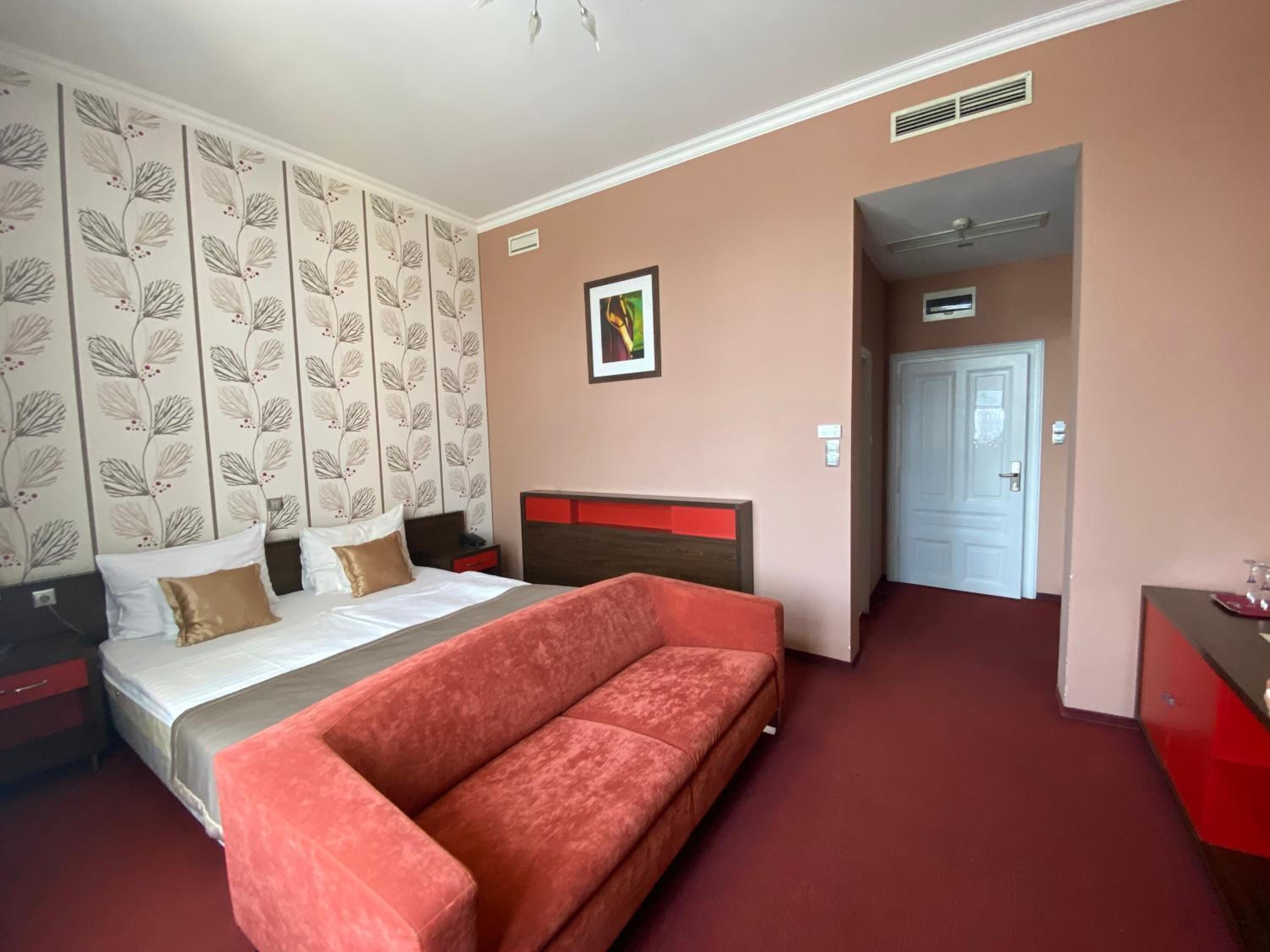 Una habitación de hotel con cama, un sofá rojo, un cuadro en la pared y una puerta que da a otra habitación.