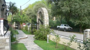 A stone pathway leading to a gated entrance with a stone archway, surrounded by lush greenery and a parked vehicle.