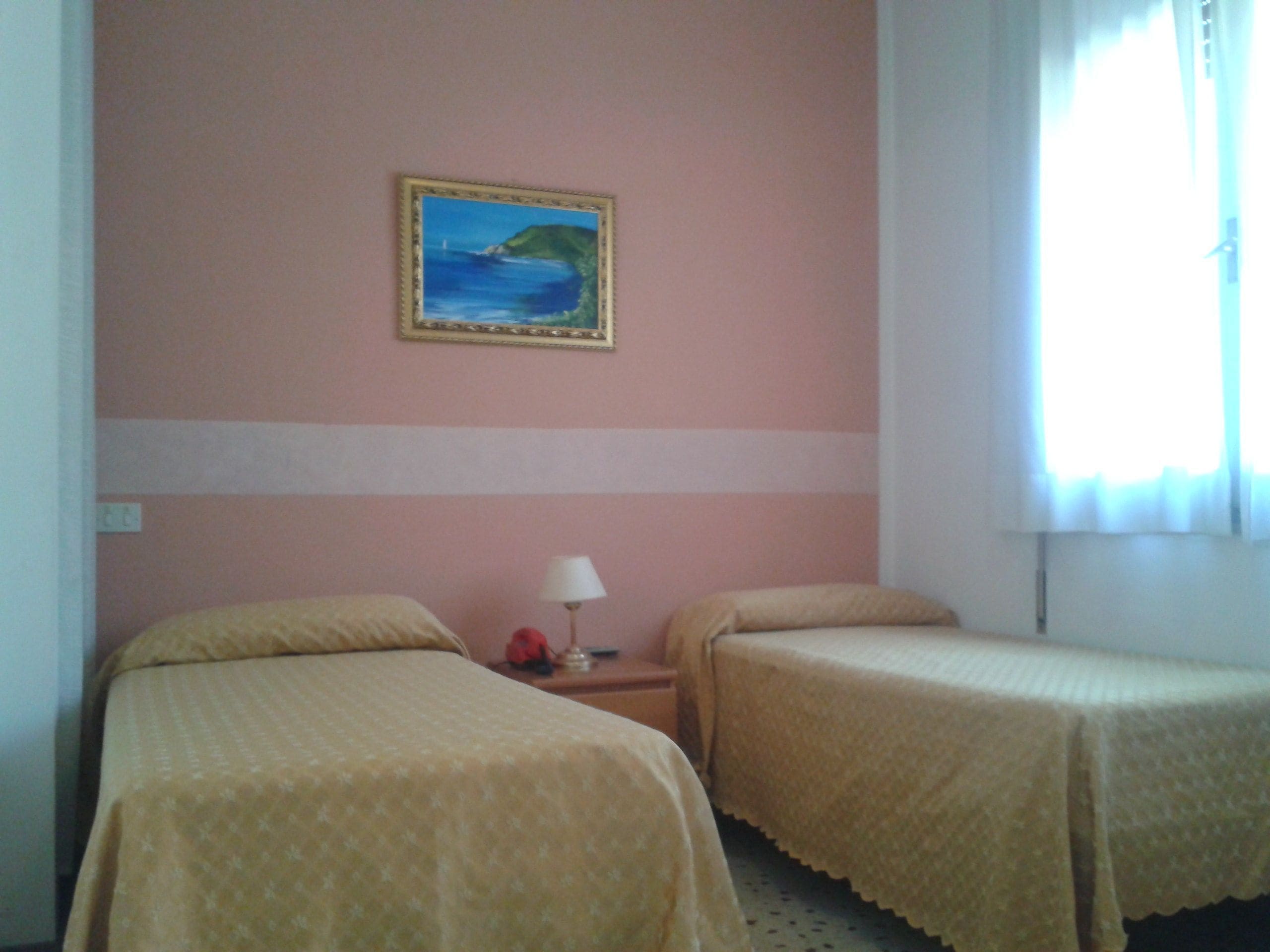 Habitación de hotel con dos camas, una mesita de noche, una lámpara y un cuadro de una escena costera en la pared.