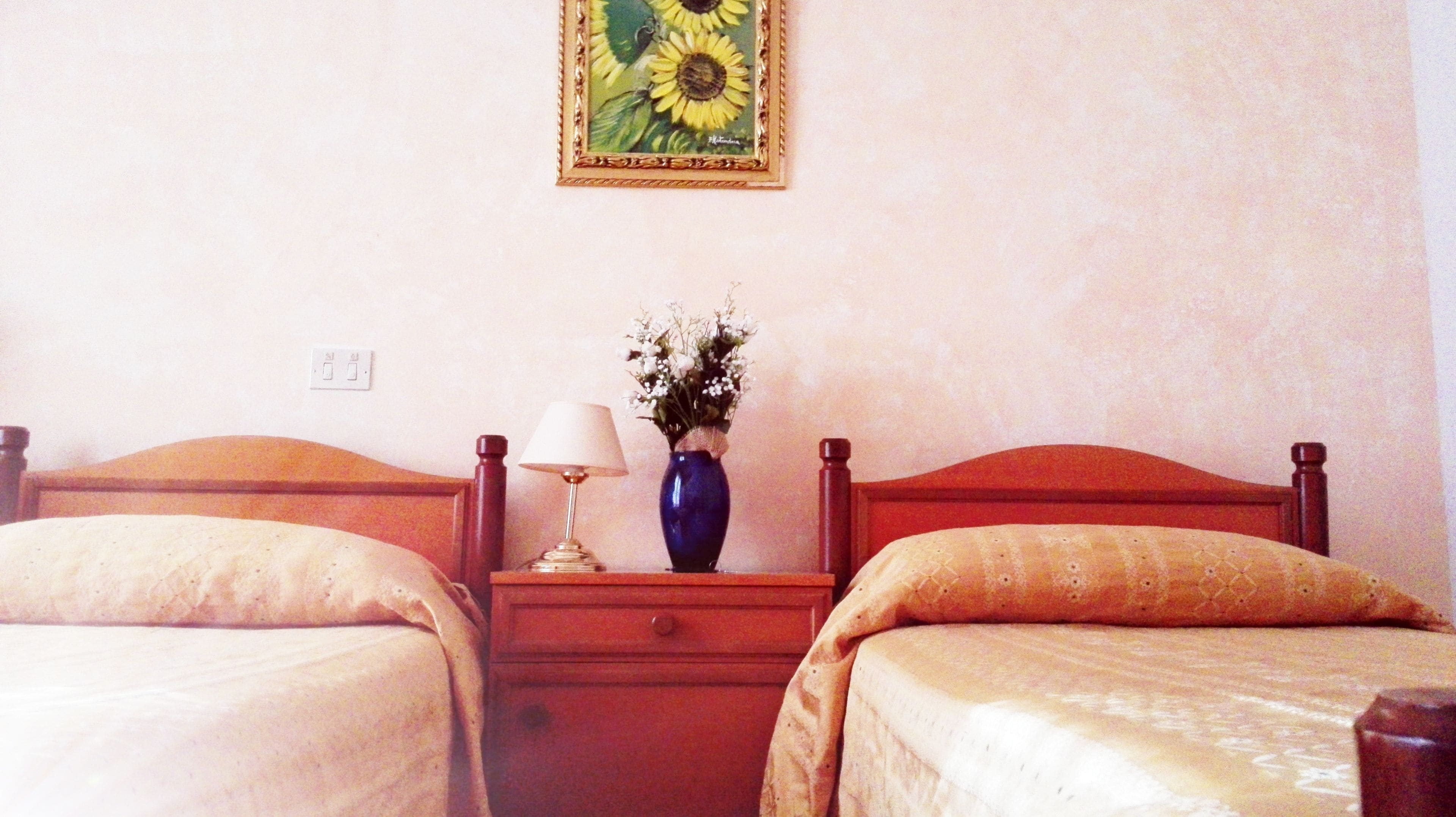 A hotel room with two beds, a nightstand with a lamp, a vase with flowers, and a painting on the wall.